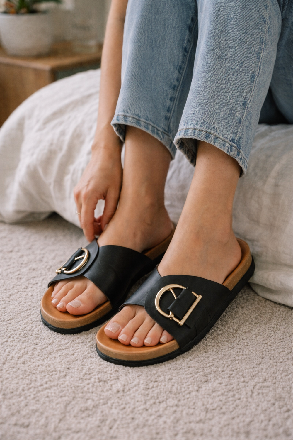 Sylvie black leather buckle slide sandals with gold-tone hardware on a woman's feet, styled with jeans.
