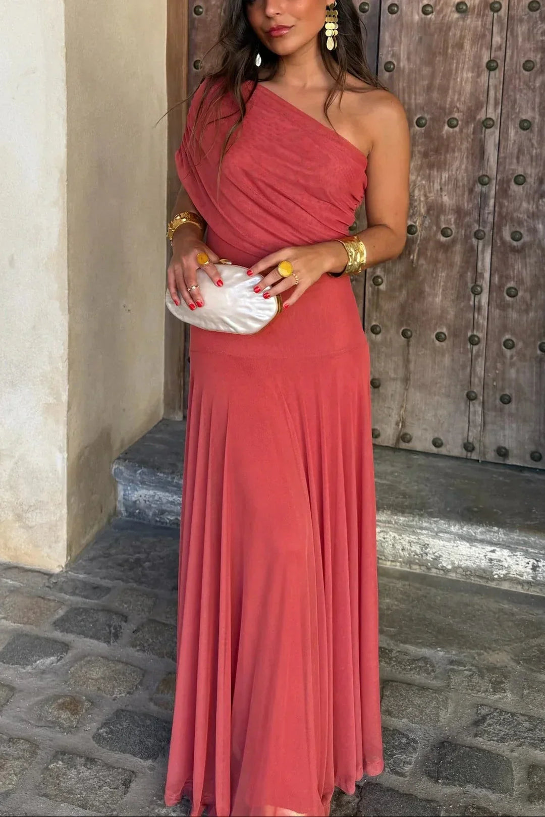 Woman in a coral Aviana one-shoulder maxi dress with draped bodice, holding a white clutch.