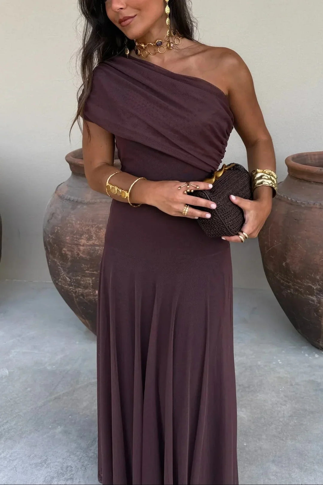 Woman in an Aviana brown one-shoulder maxi dress with draped bodice, holding a clutch, wearing gold jewelry.