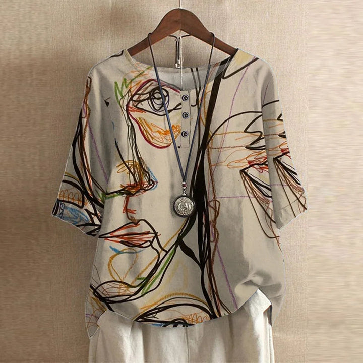 Beige short-sleeve blouse with a colorful abstract face print, button-front neckline, and a relaxed, draped fit.
