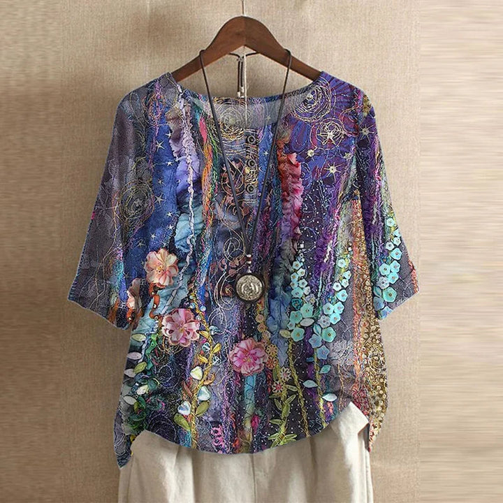 Relaxed-fit blouse with a vibrant purple floral print, round neckline, and three-quarter sleeves on a wooden hanger.