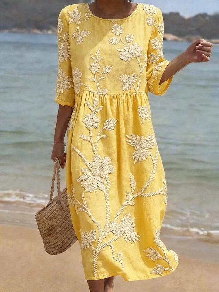 Yellow midi dress with white embroidered vintage floral design, three-quarter sleeves, and a relaxed fit, worn on a beach.