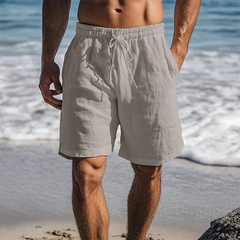 Men's light gray linen drawstring shorts with side pockets, worn on a beach.