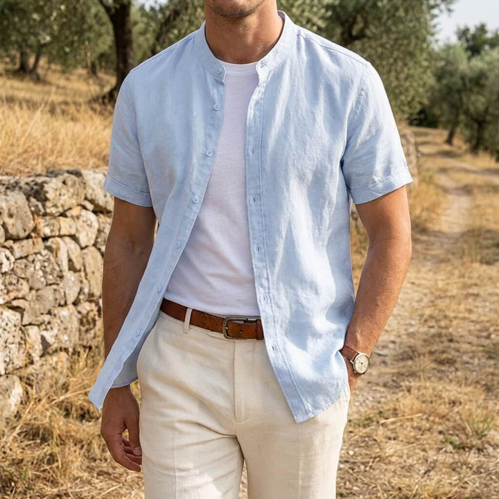 Men's light blue short sleeve linen shirt with stand collar, worn open over a white tee.