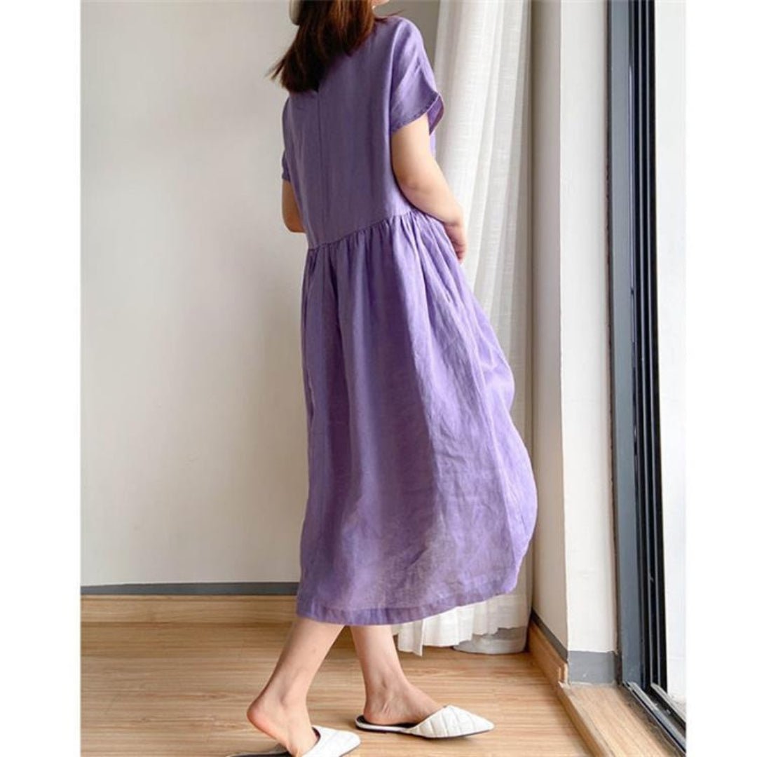 Woman in a relaxed purple linen short sleeve midi dress with a gathered waist, standing by a window.