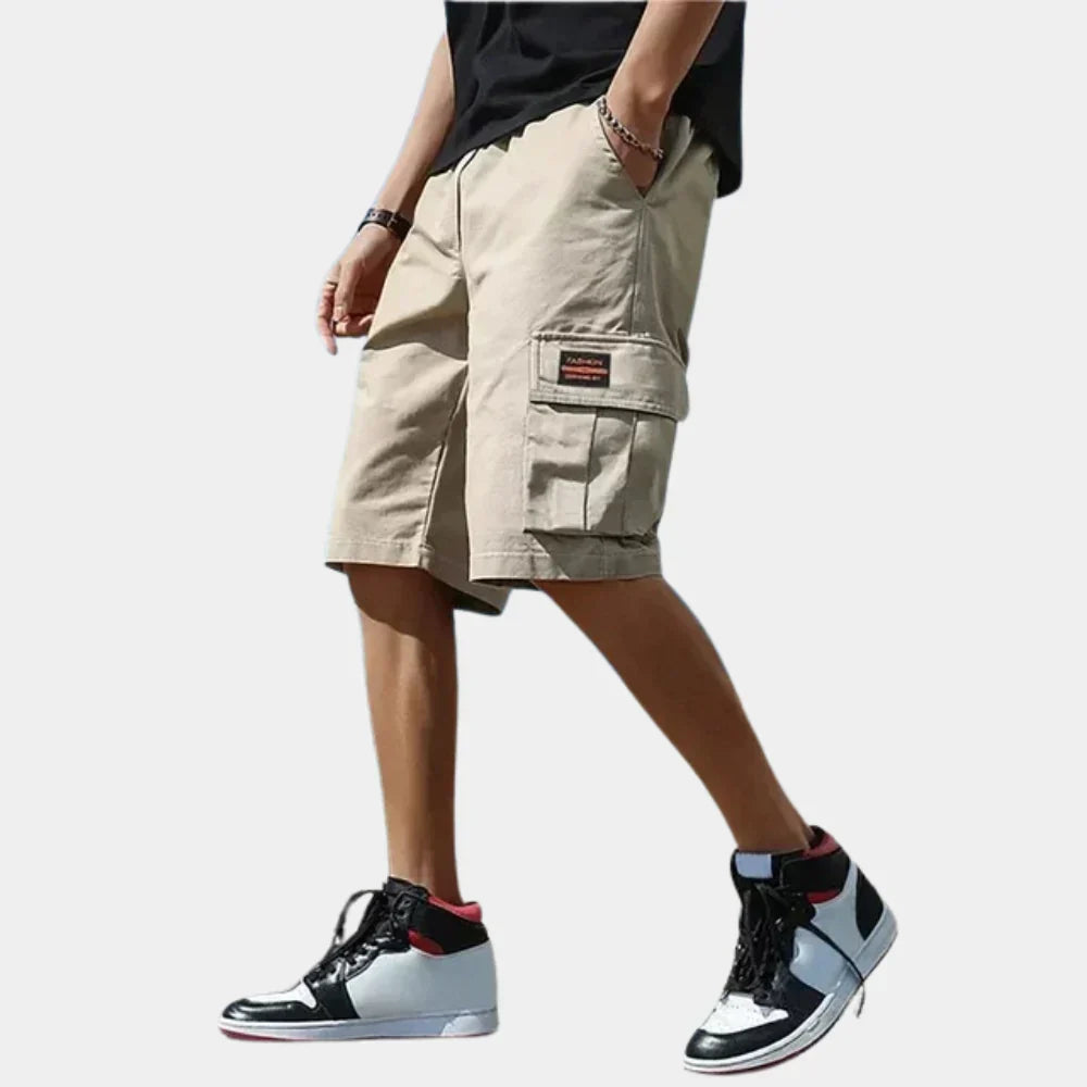 Beige men's cargo shorts with a drawstring waist, loose fit, and side utility pockets, styled with high-top sneakers.