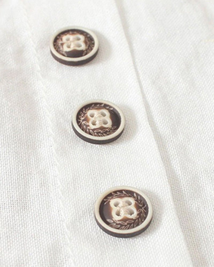 Close-up of white linen fabric featuring decorative brown buttons with intricate leaf-patterned detailing.