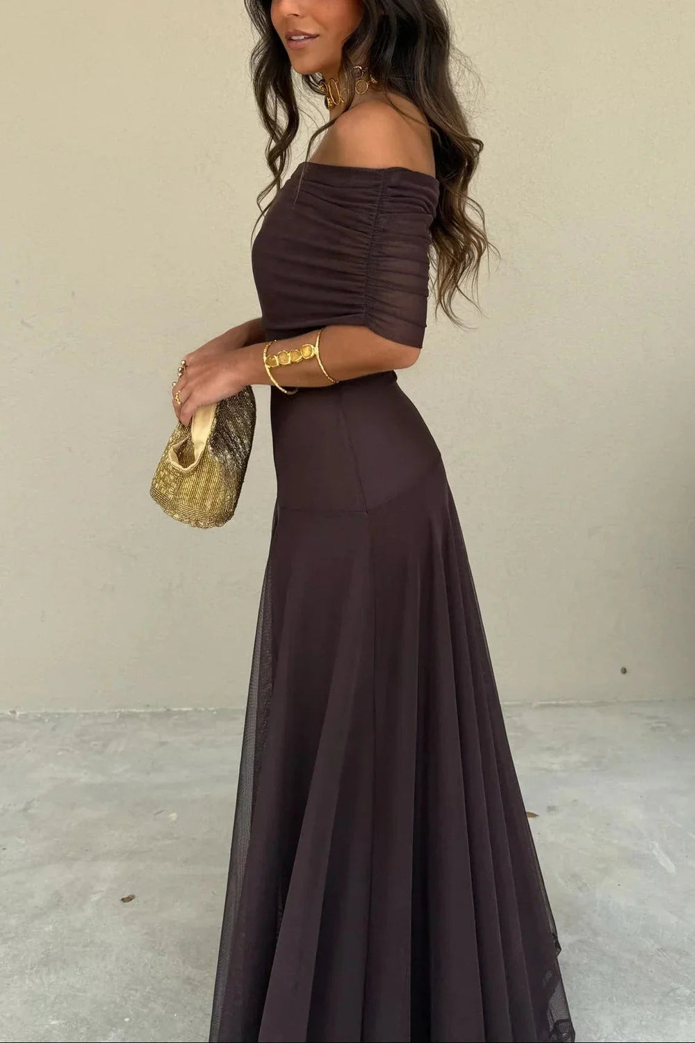 Nora women's one-shoulder pleated maxi dress in rich brown, flowing gracefully with a gold clutch.